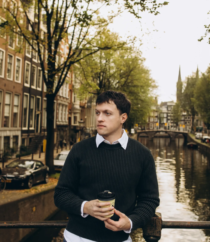 Man holding a coffee at a canal railing with a bridge behind, Amsterdam