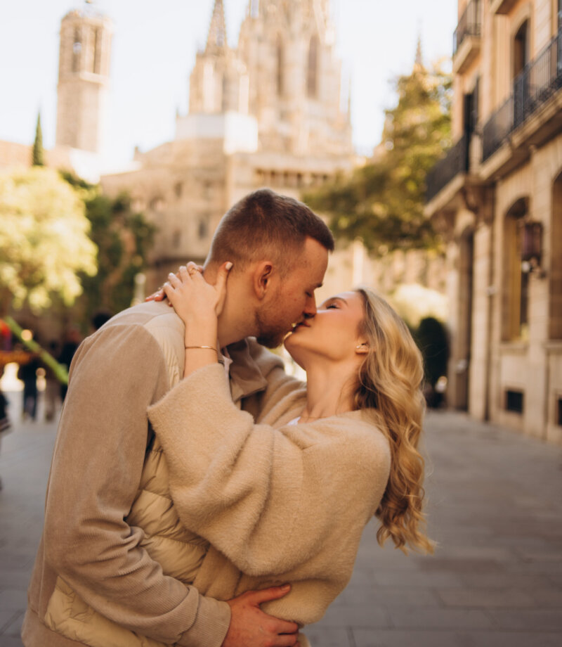 Couple in beige knitwear kissing on a street with the spires of Barcelona Cathedral behind