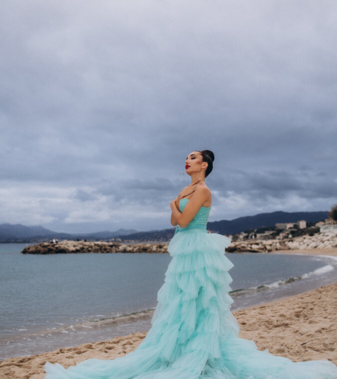 Woman in a tiered turquoise tulle gown standing on the beach with the Bay of Cannes behind