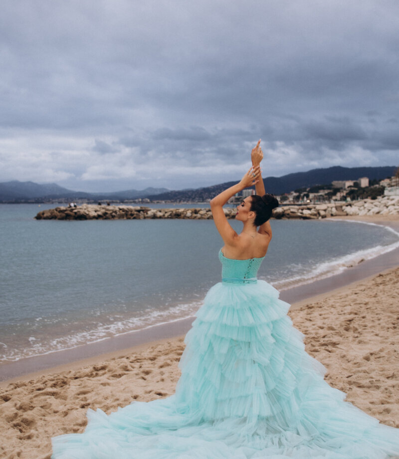 Woman with arms raised in a turquoise tulle gown on a Cannes beach under a stormy sky