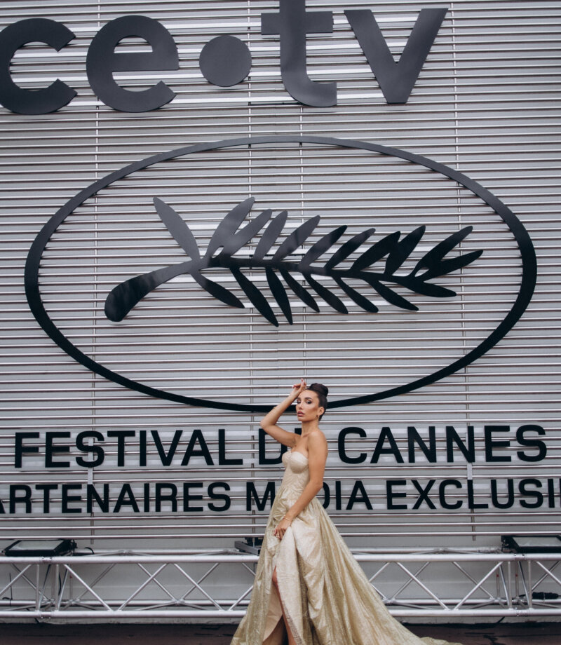 Woman in a gold gown posing before the Festival de Cannes palm logo backdrop