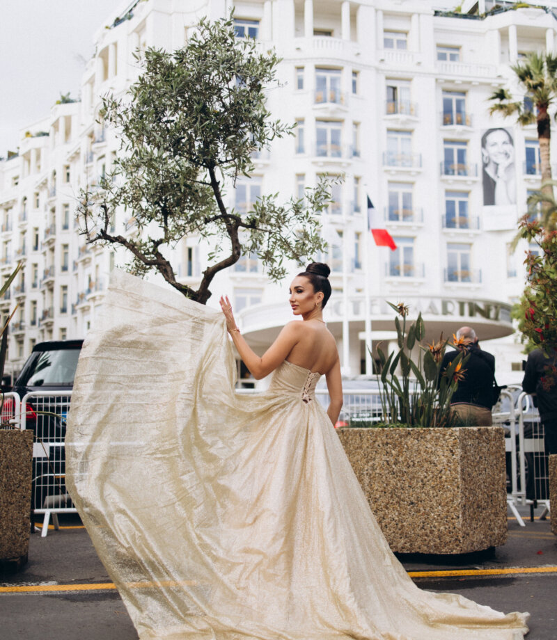 Woman tossing the long train of a gold gown in front of Hotel Barrière Le Majestic, Cannes
