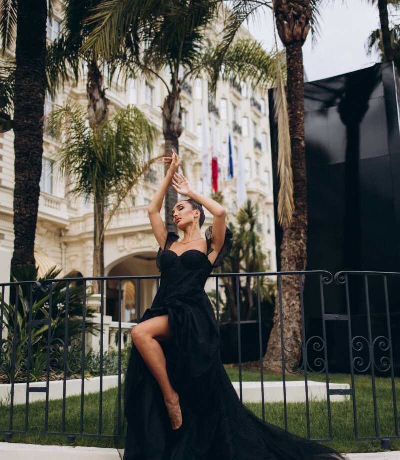 Woman in a black slit gown posing among palm trees in front of a Belle Époque hotel on La Croisette, Cannes