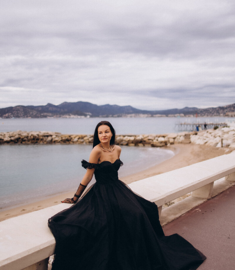 Woman in a black gown seated on a seafront bench on La Croisette with the Lérins coast behind, Cannes