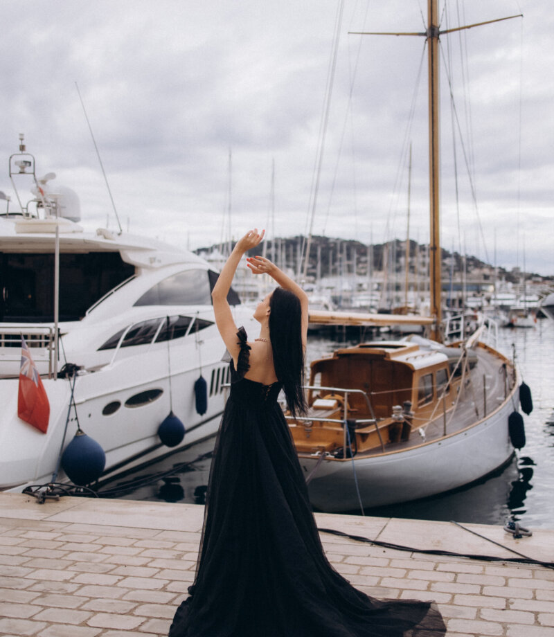 Dark-haired woman in a black gown posing by a classic wooden sailboat in the Vieux Port of Cannes