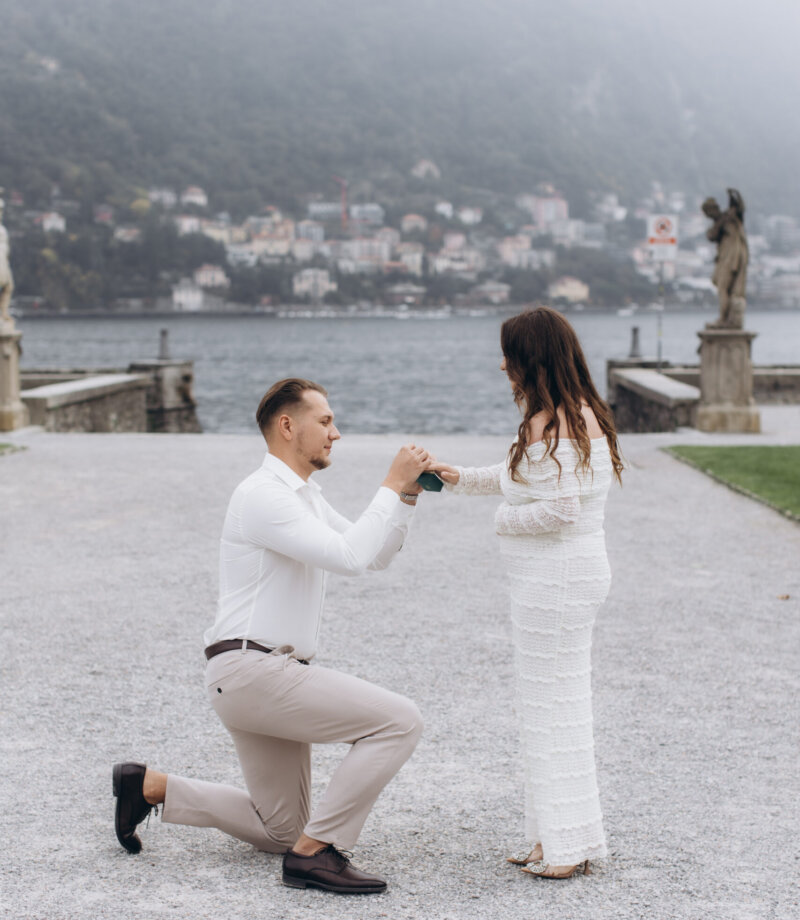 Man kneeling to hold her hands as she accepts the ring by a statue on the Villa Olmo lakefront, Lake Como