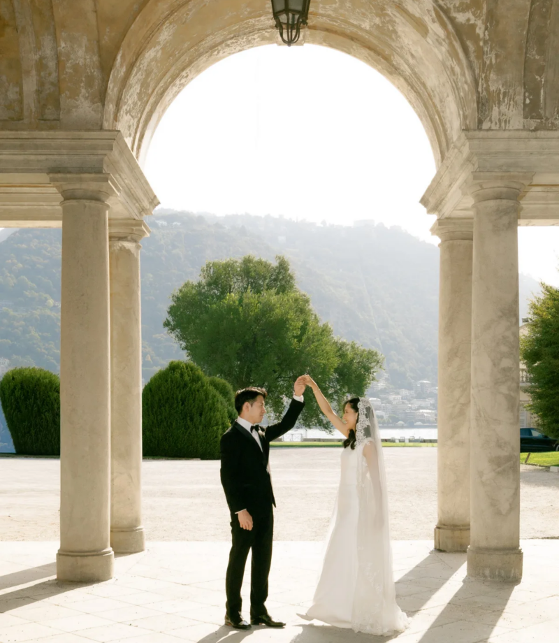 Groom twirling the bride under a stone arcade with a lantern and the lake behind, Villa Olmo, Lake Como
