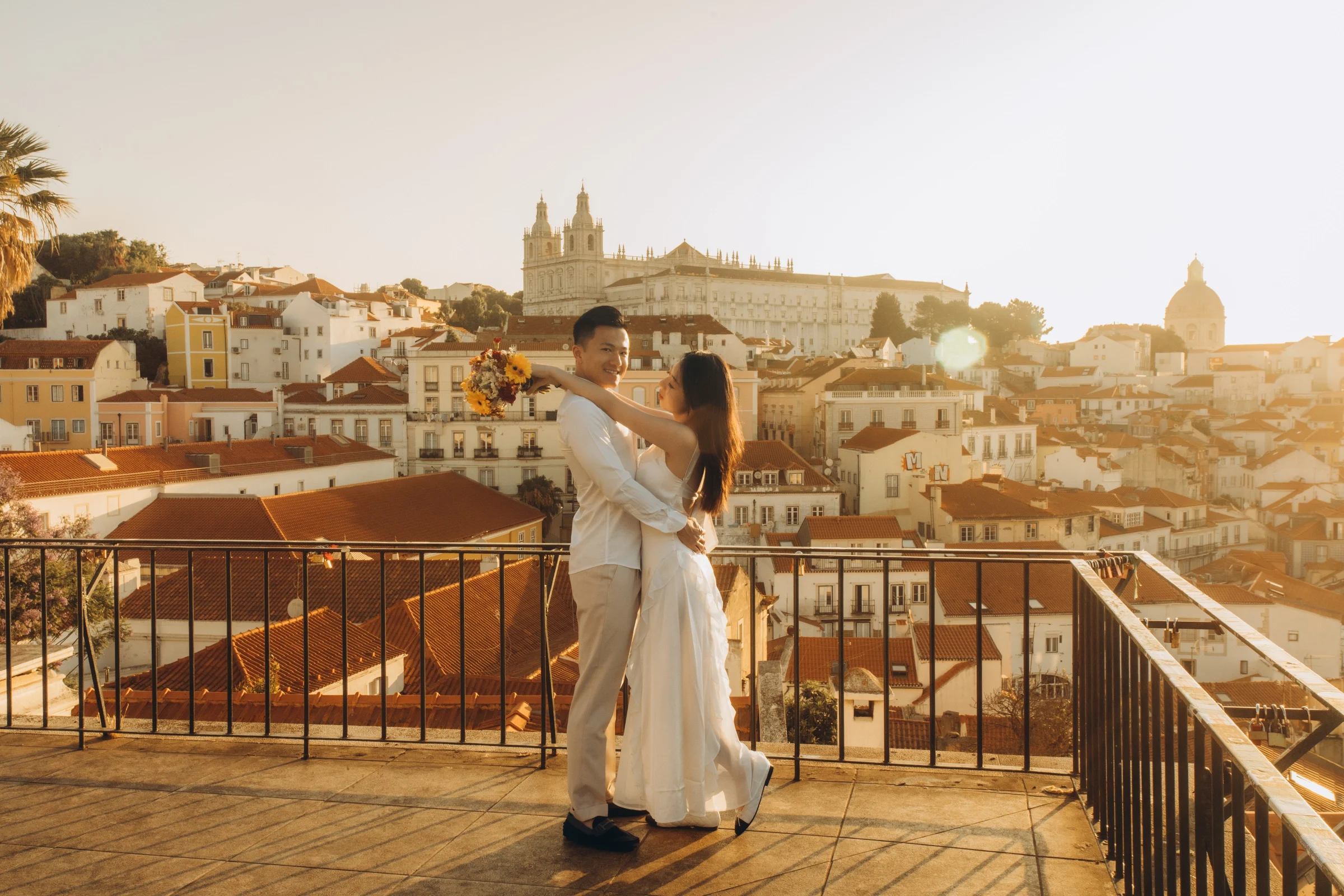 Couple embracing at an Alfama viewpoint at sunrise, the rooftops and domes of Lisbon glowing behind them