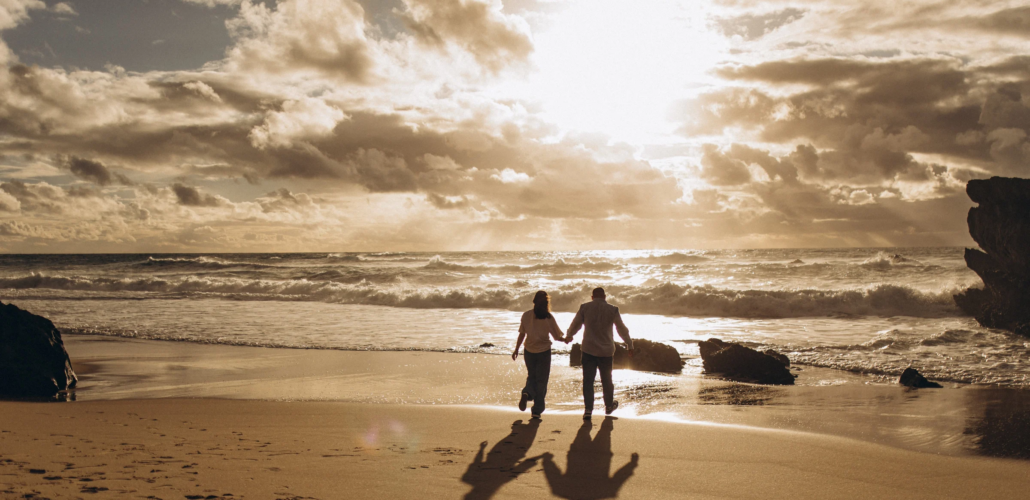 Couple walking hand in hand on an Atlantic beach under a dramatic golden sunset sky near Lisbon