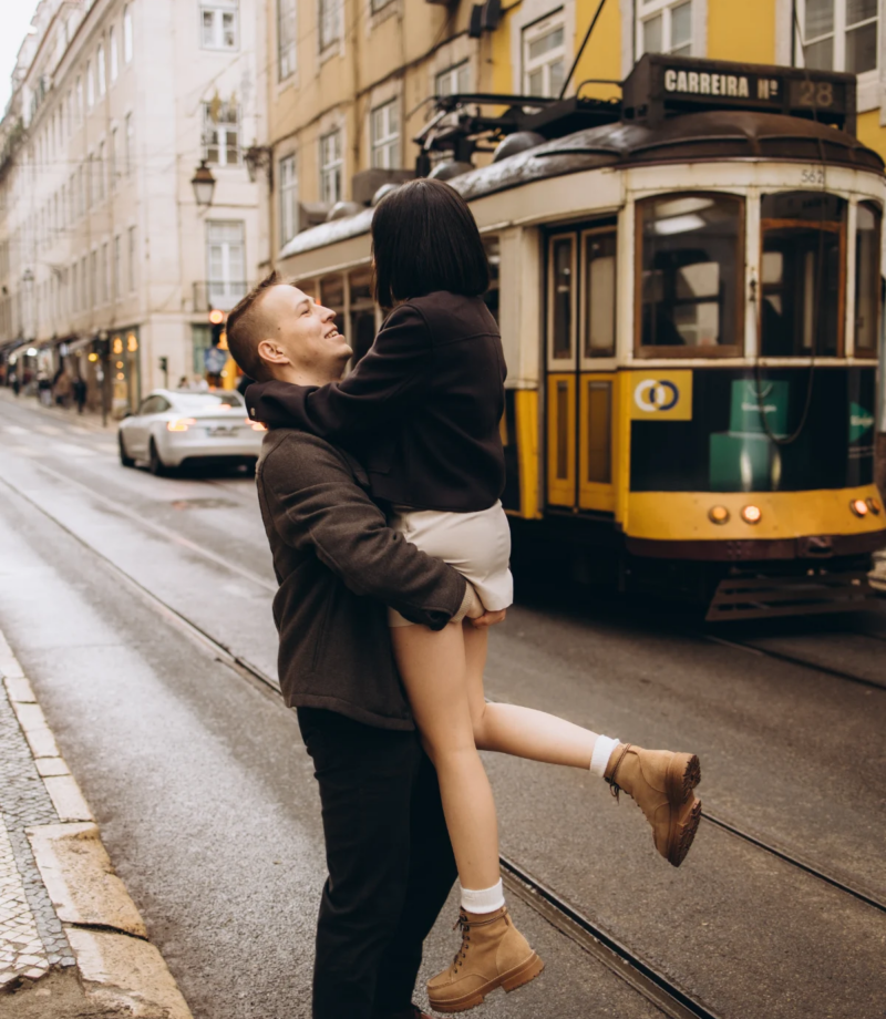 Man lifting his partner beside a yellow tram on a Lisbon street