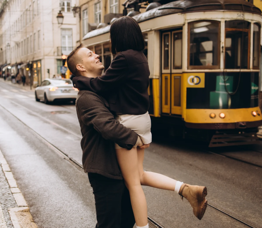 Man lifting his partner beside a yellow tram on a Lisbon street