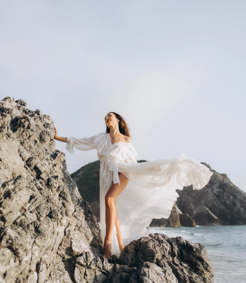 Woman in a white dress on the rocks at Praia da Adraga, the dress lifting in the wind, near Lisbon
