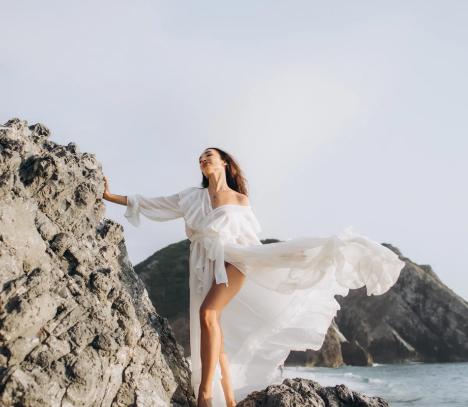 Woman in a white dress on the rocks at Praia da Adraga, the dress lifting in the wind, near Lisbon