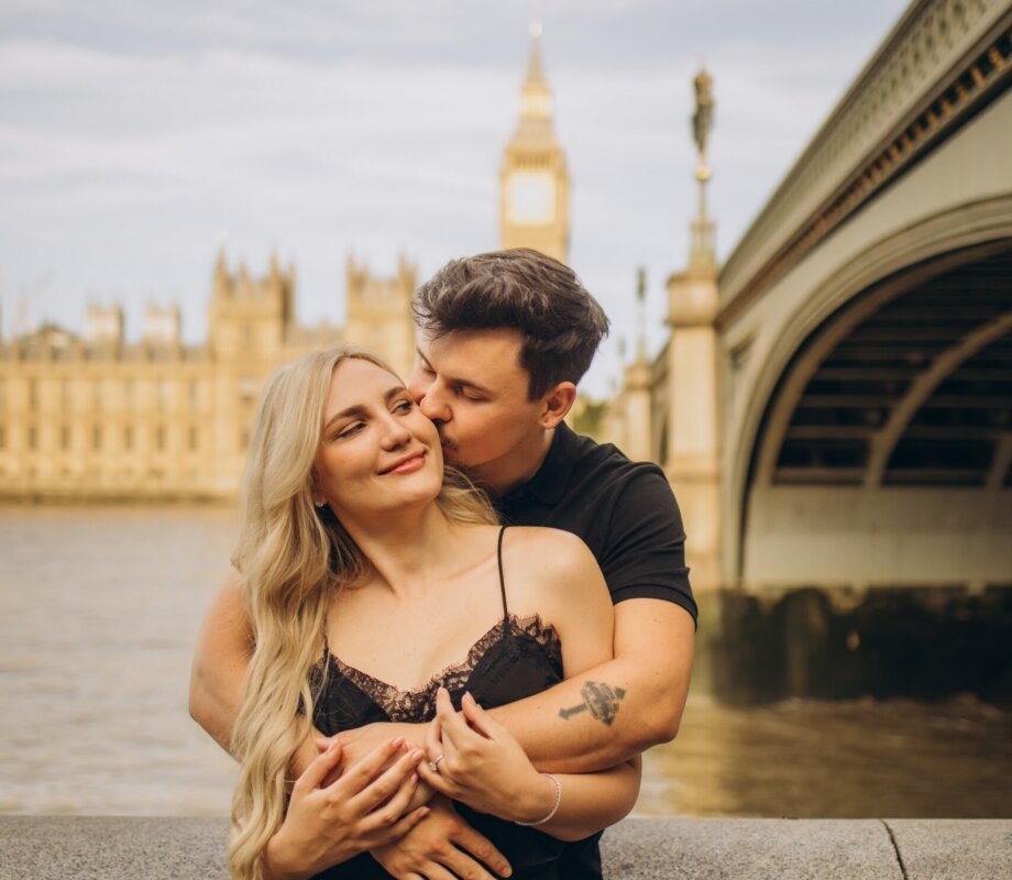 Man kissing his partner's cheek by Westminster Bridge in golden light, London