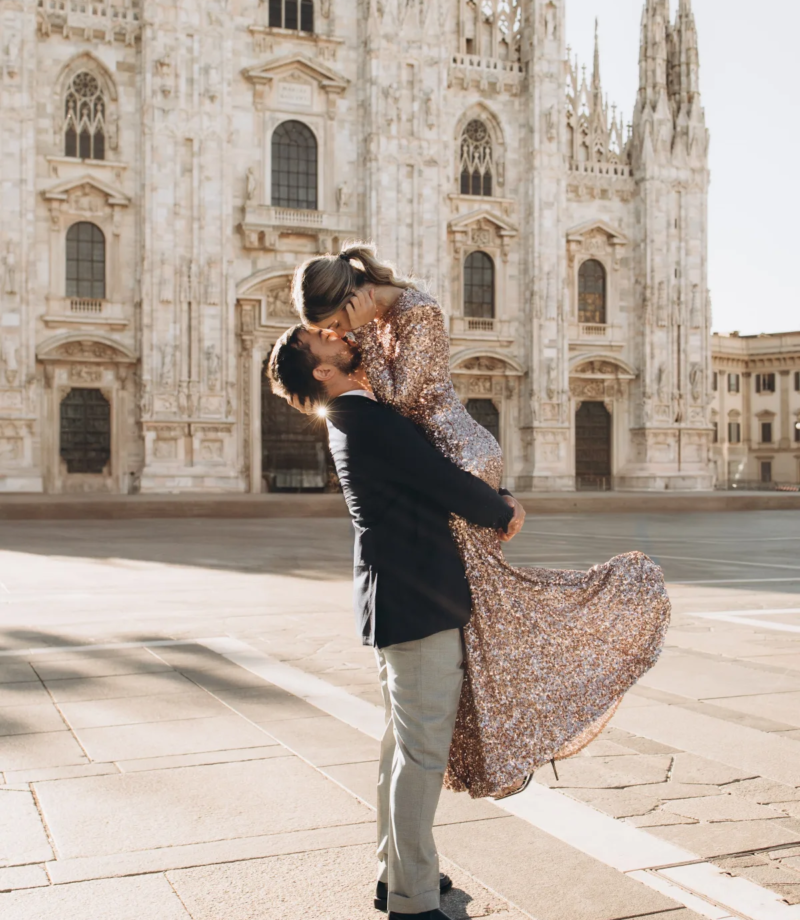 Man lifting a woman in a sequin gown off the ground for a kiss in front of the Duomo, Milan