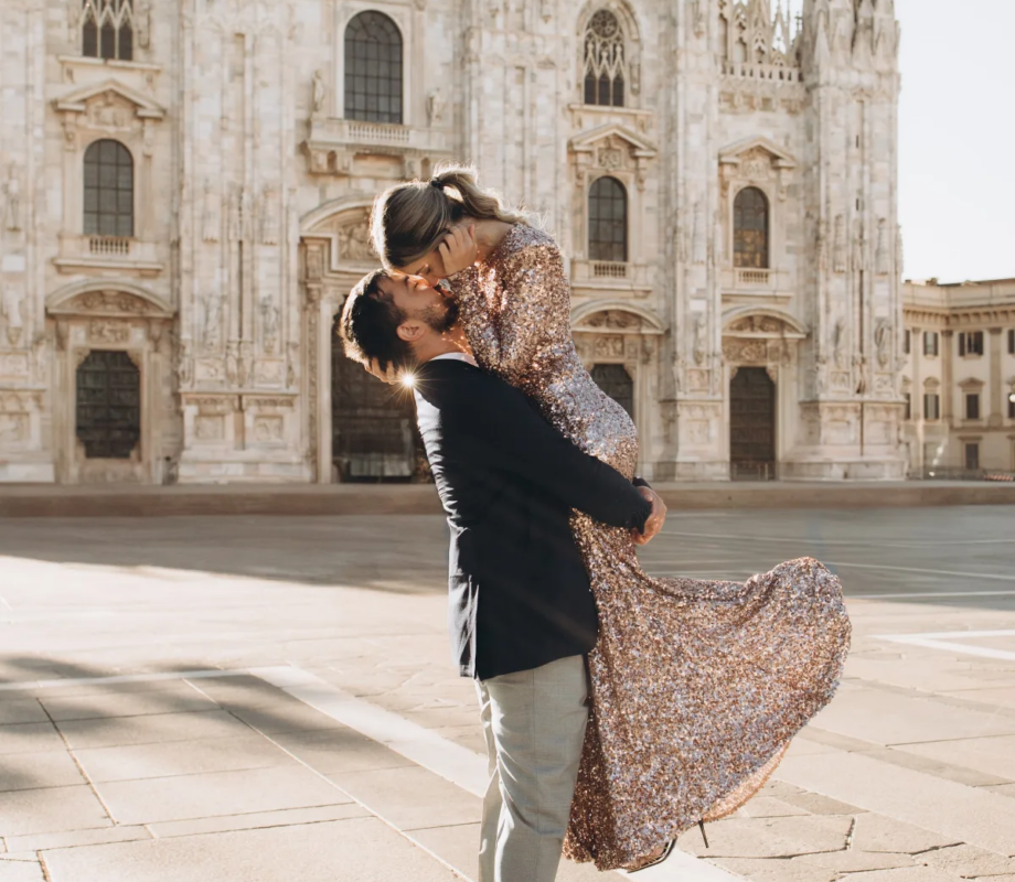 Man lifting a woman in a sequin gown off the ground for a kiss in front of the Duomo, Milan