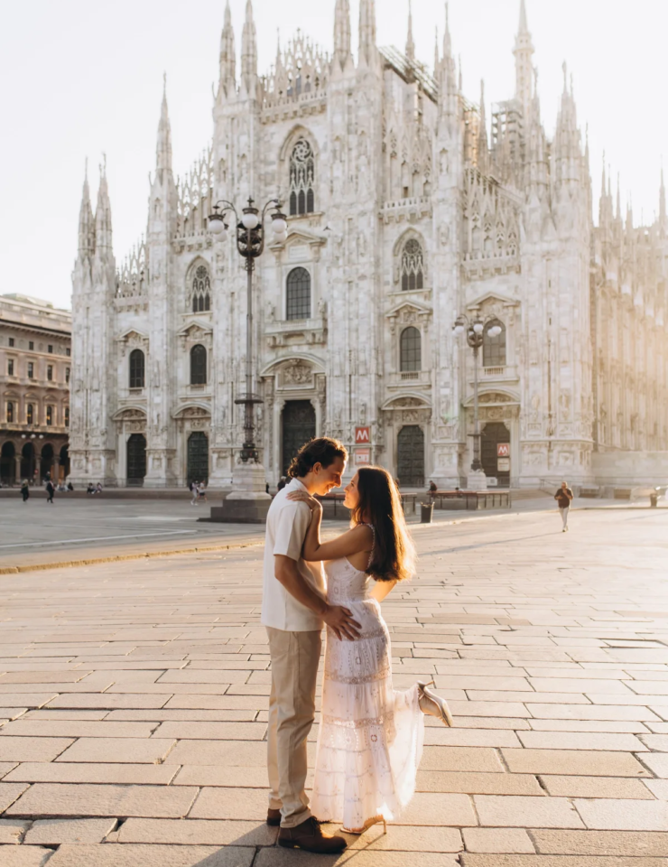 Woman in a white lace dress lifting one foot as she kisses her partner in the open Piazza del Duomo, Milan