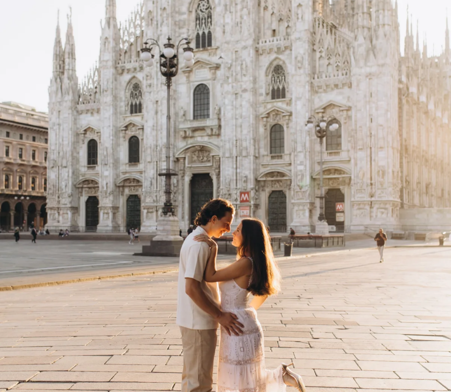 Woman in a white lace dress lifting one foot as she kisses her partner in the open Piazza del Duomo, Milan