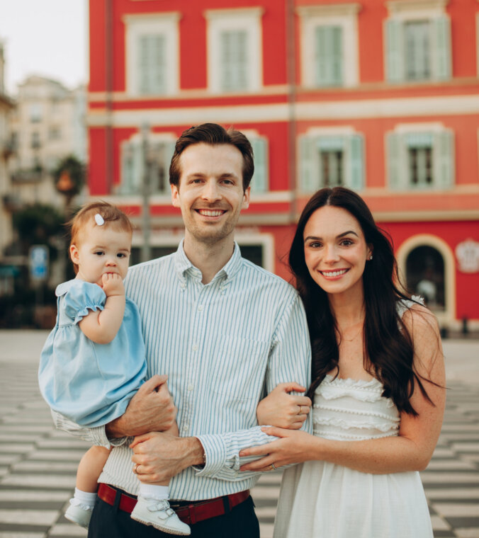 Family of three posing in front of the red Italianate buildings of Place Masséna, Nice