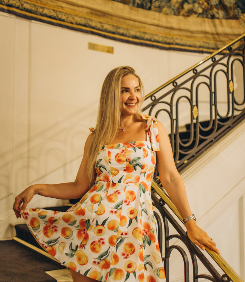 Blonde woman in a floral sundress posing on a curved wrought-iron staircase in Hotel Negresco, Nice