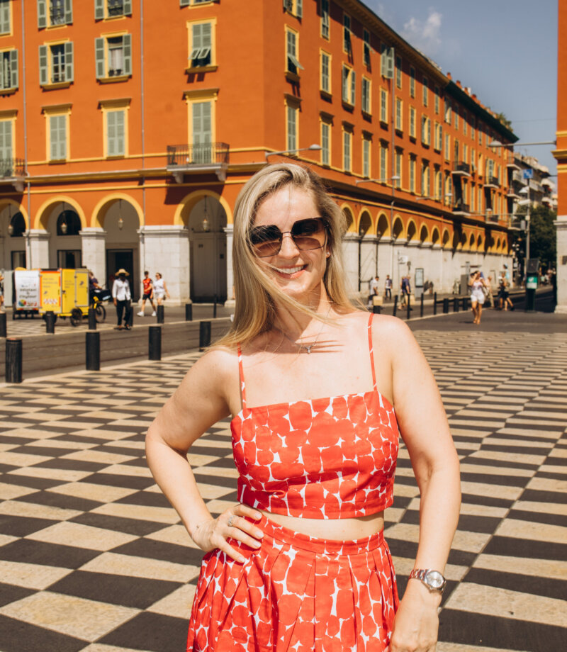 Blonde woman in a red summer dress standing on the checkerboard paving of Place Masséna, Nice
