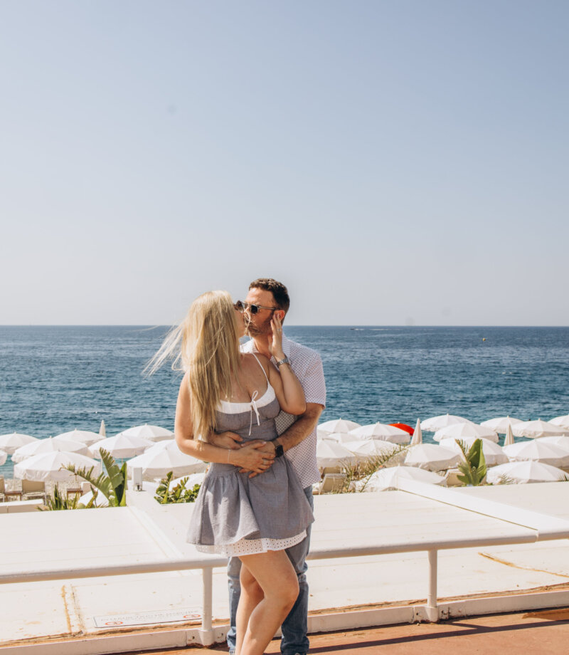 Couple hugging by the railing above a beach of white parasols on the Promenade des Anglais, Nice