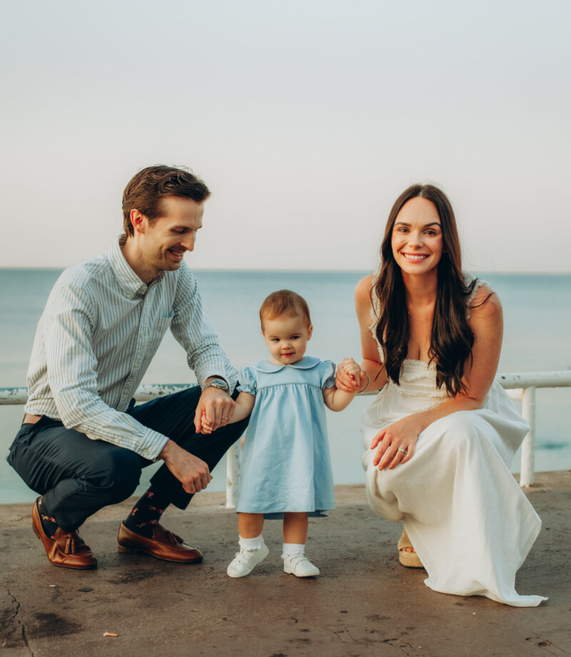 Parents crouching beside their toddler in a blue dress by the sea on the Promenade des Anglais, Nice