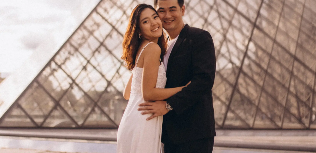 Couple in a white dress and black suit embracing at the Louvre glass pyramid at golden hour, Paris