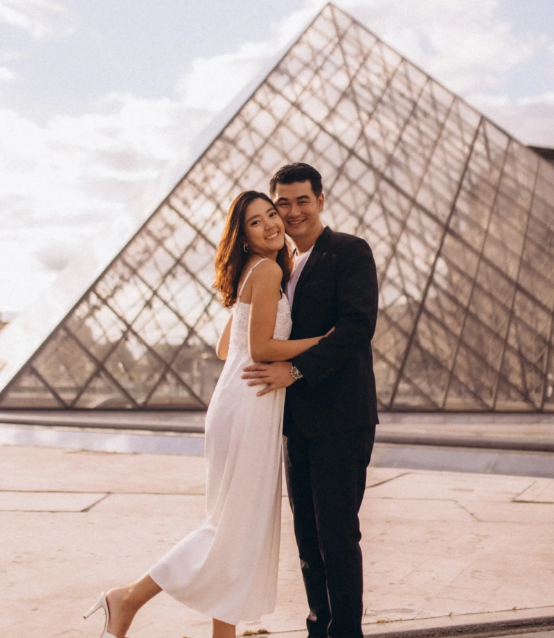 Couple in a white dress and black suit embracing at the Louvre glass pyramid at golden hour, Paris