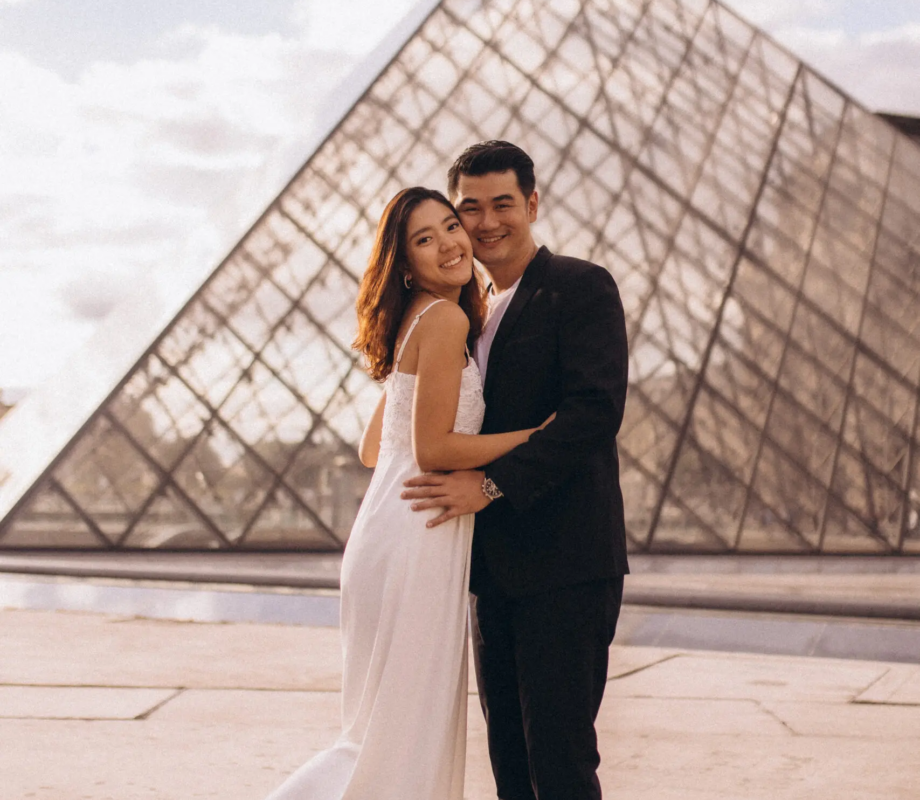 Couple in a white dress and black suit embracing at the Louvre glass pyramid at golden hour, Paris