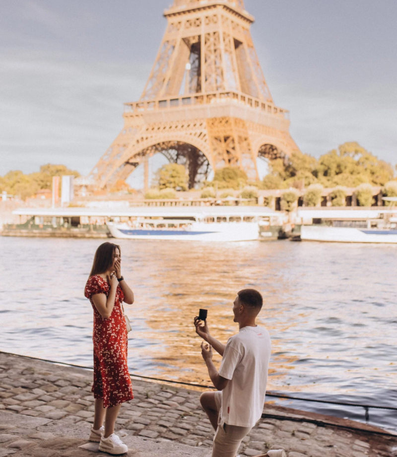 Man on one knee proposing to a woman in a red dress by the Seine with the Eiffel Tower behind, Paris