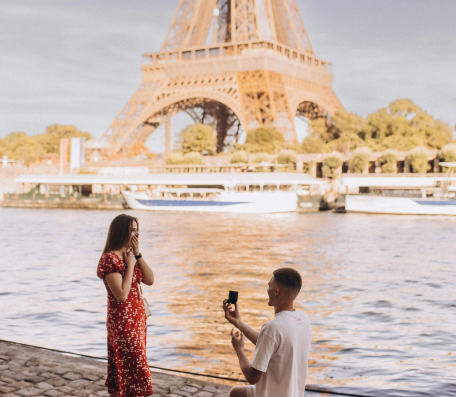 Man on one knee proposing to a woman in a red dress by the Seine with the Eiffel Tower behind, Paris
