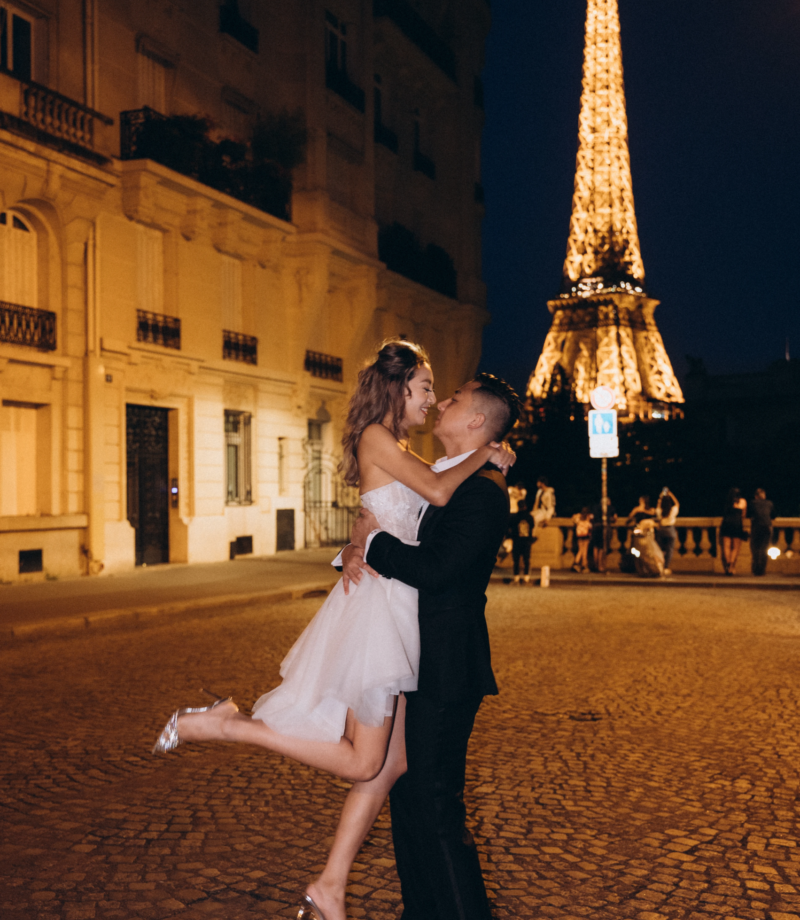 Groom lifting the bride on a cobbled street at night with the sparkling Eiffel Tower behind, Paris