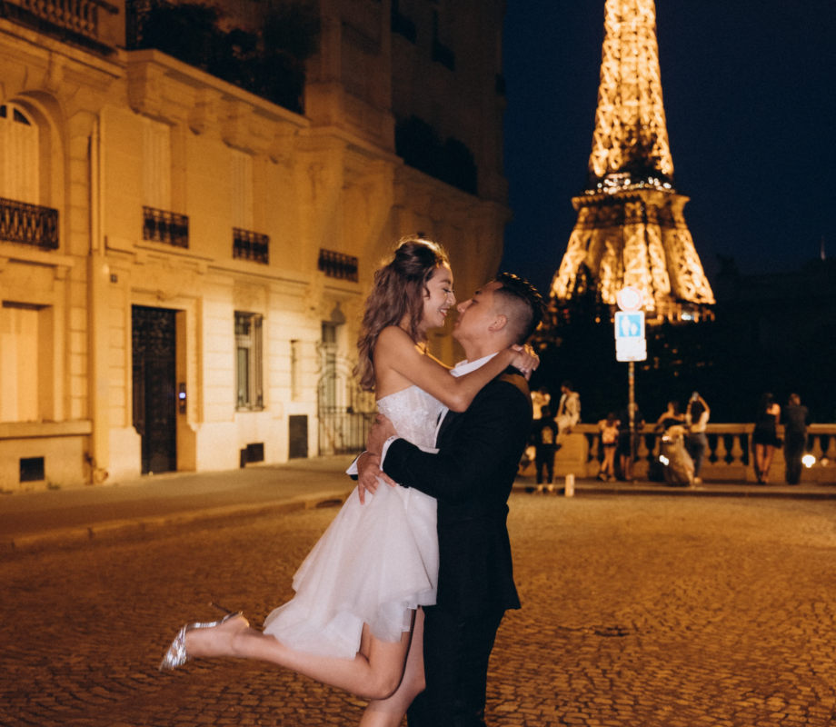 Groom lifting the bride on a cobbled street at night with the sparkling Eiffel Tower behind, Paris