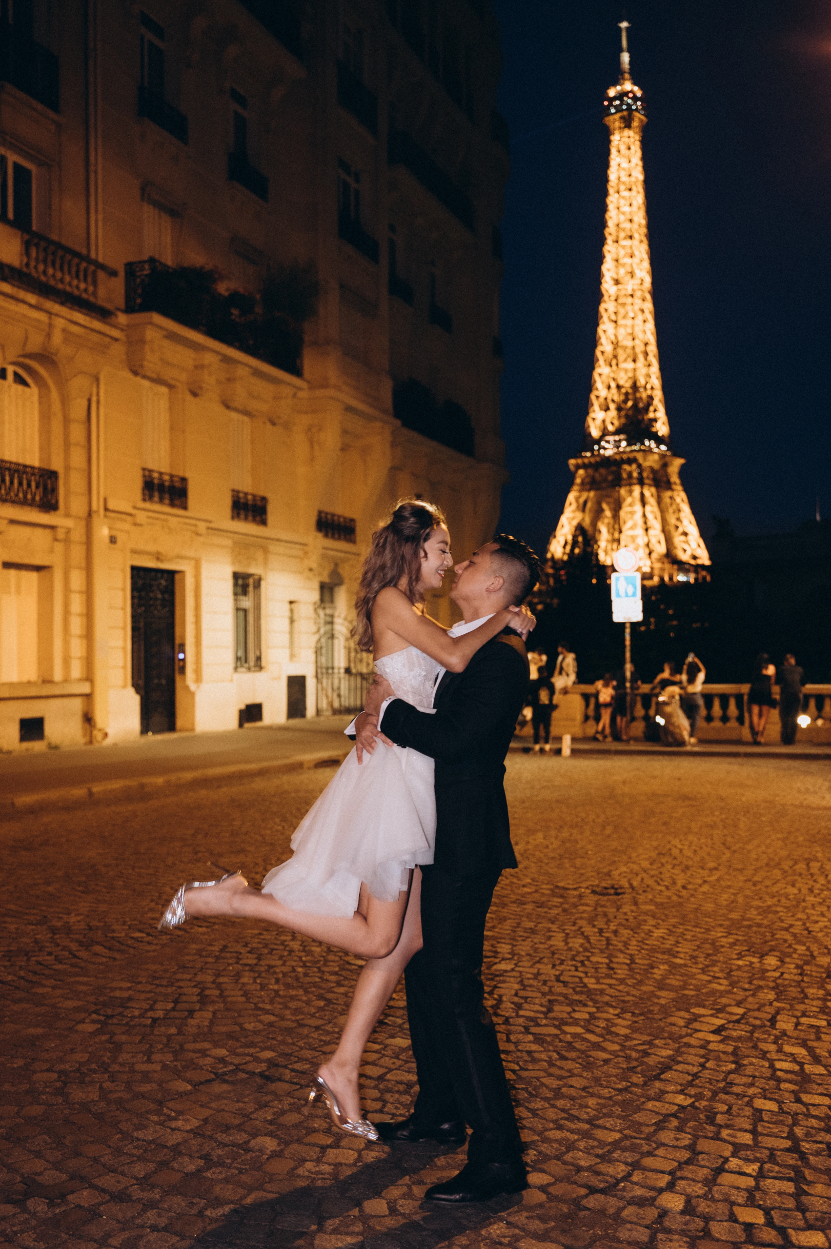 Groom lifting the bride on a cobbled street at night with the sparkling Eiffel Tower behind, Paris