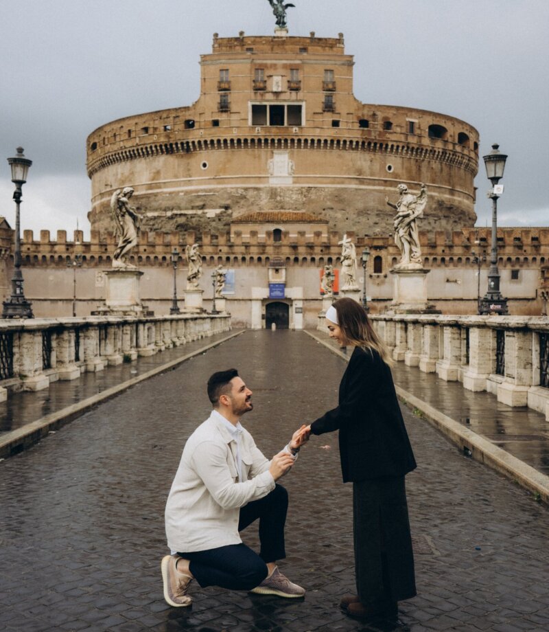 Man on one knee proposing on Ponte Sant'Angelo with Castel Sant'Angelo behind, Rome