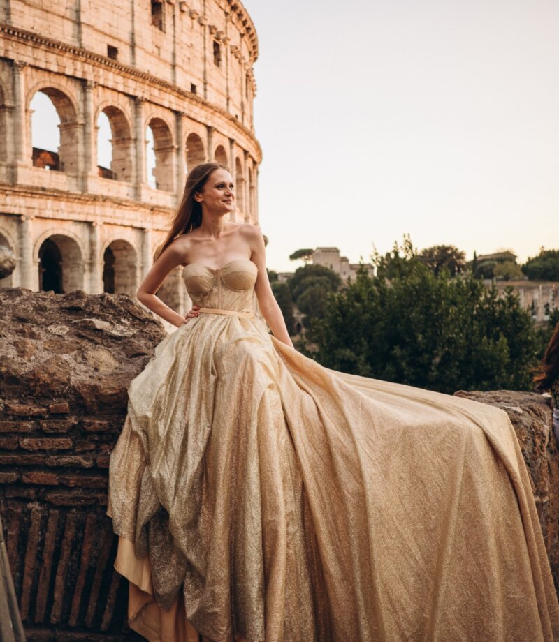 Gold flying dress with its train spread over ancient stones by the Colosseum, Rome
