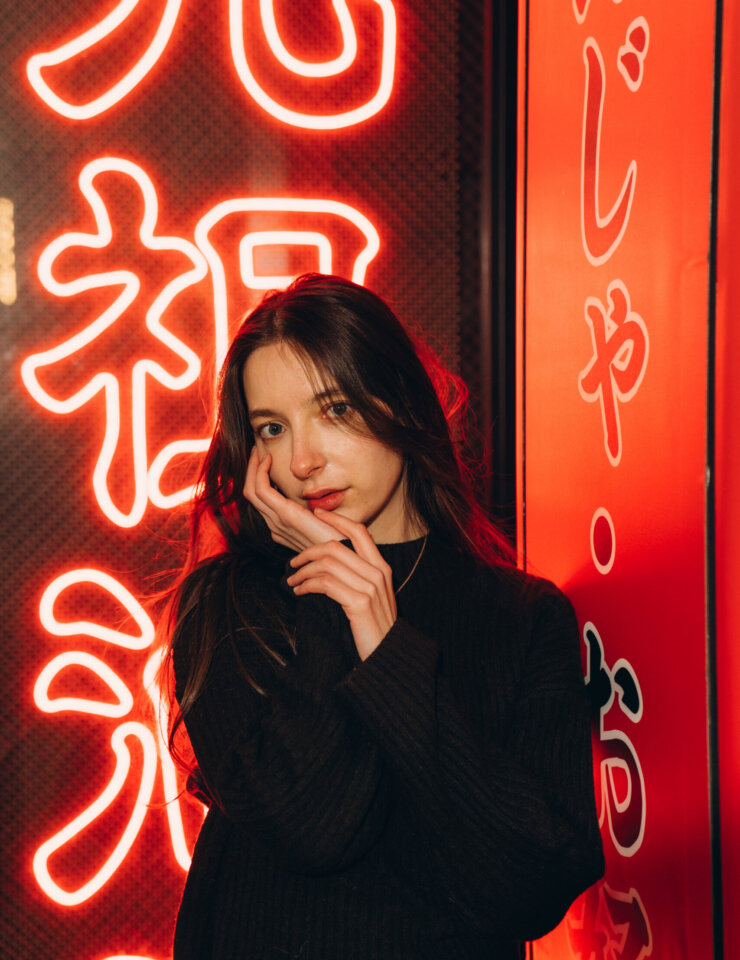 Young woman in a black sweater touching her cheek in front of a red neon kanji sign in Shibuya, Tokyo