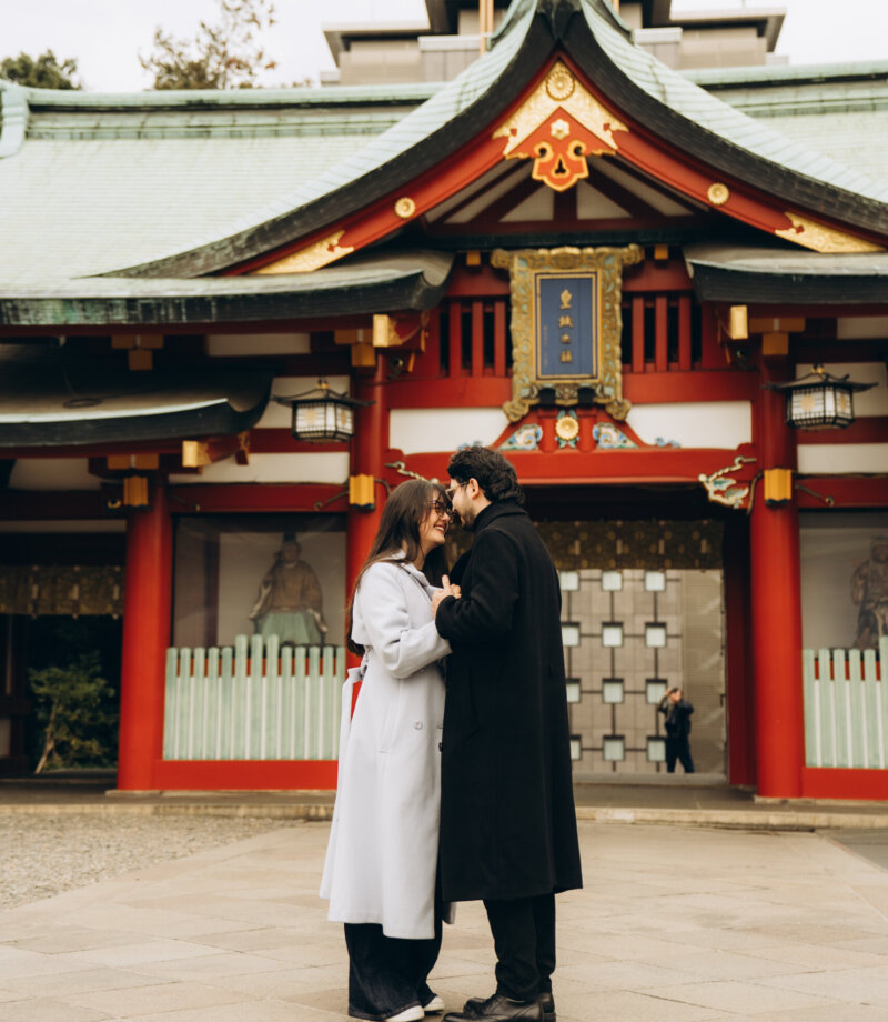 Couple in black and pale grey coats embracing in front of the vermilion main hall of Hie Shrine, Tokyo
