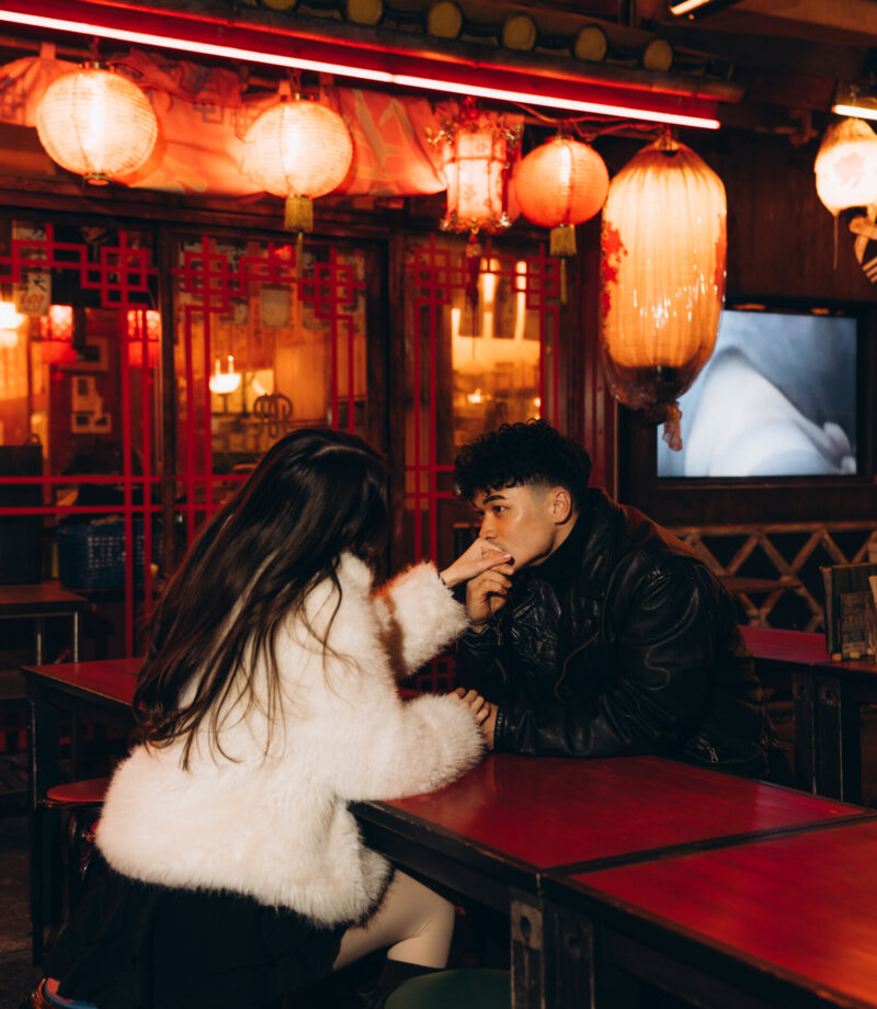 Couple sitting at a red lacquered table in a lantern-lit Tokyo food hall, leaning in close